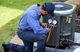 Technician in blue uniform kneeling while checking HVAC unit gauges outdoors.