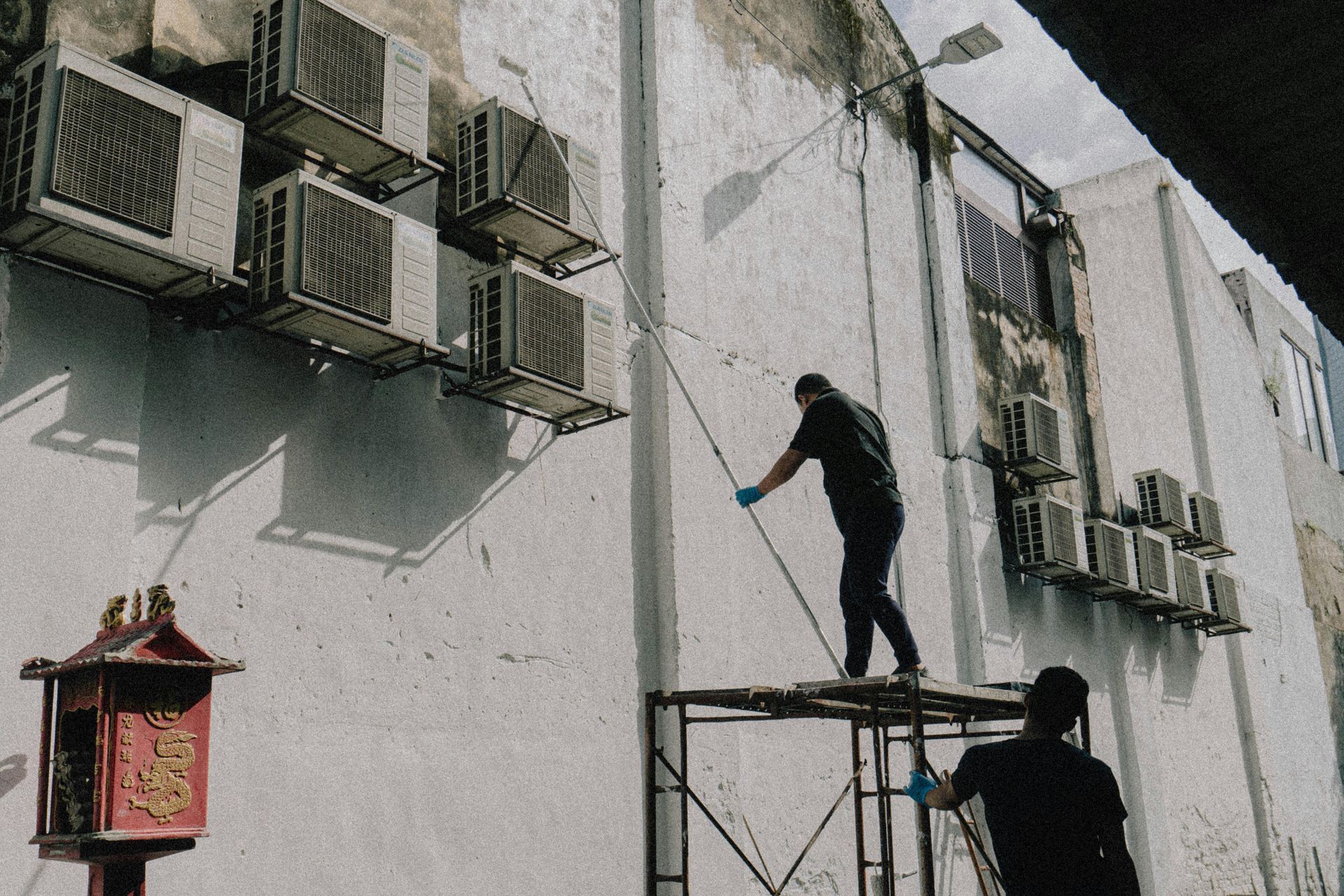 Two people spraying a white building. Scaffolding is set up. Many AC units are mounted on the wall.