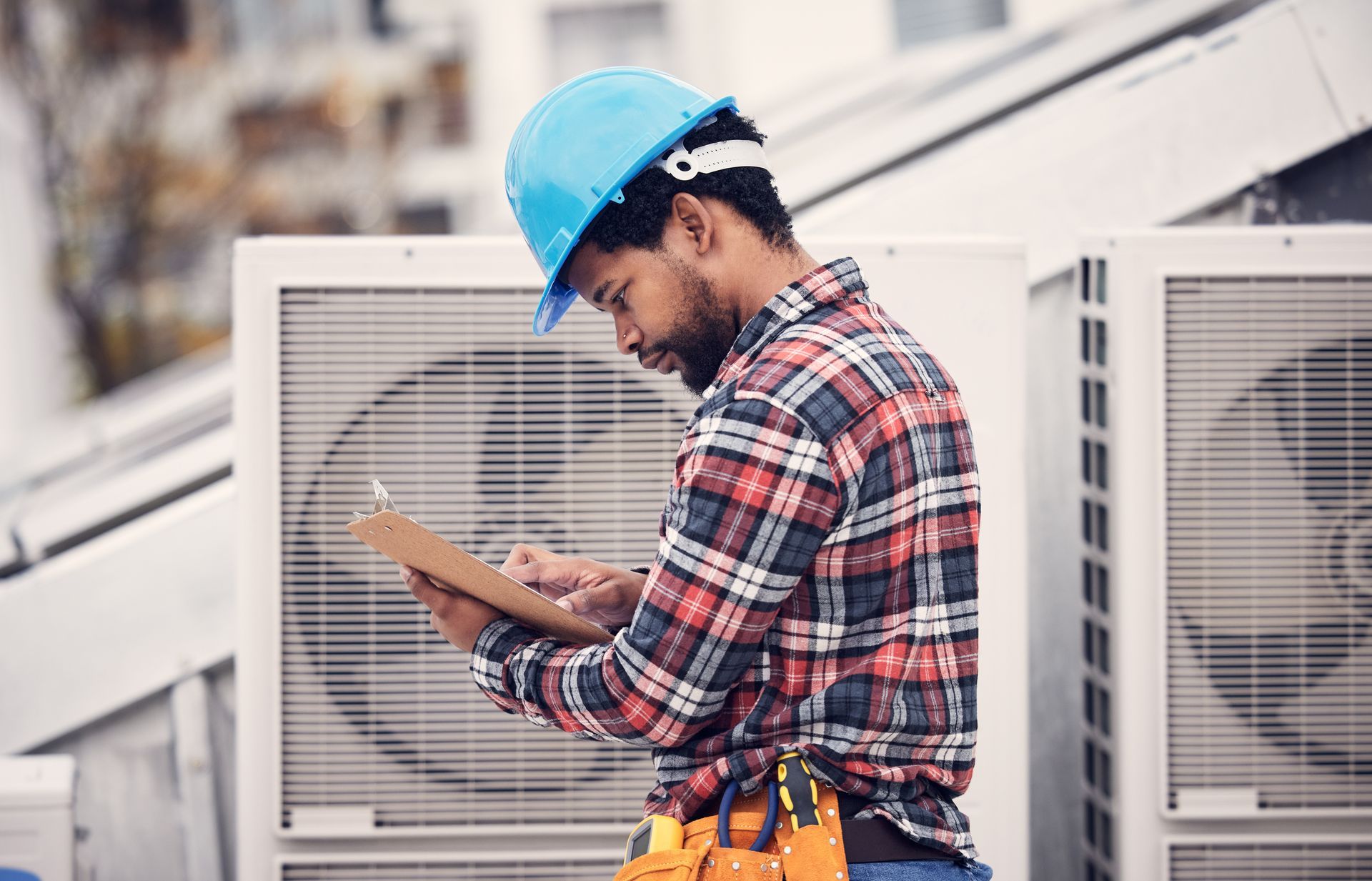 HVAC technician in hard hat and plaid shirt inspects rooftop air conditioning units, holding a clipboard.