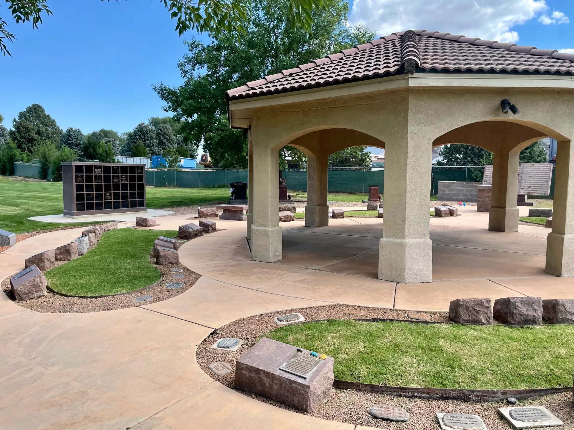 Gazebo with tile roof in park.  Pathways with rocks and greenery lead to memorial structures. Sunny day.
