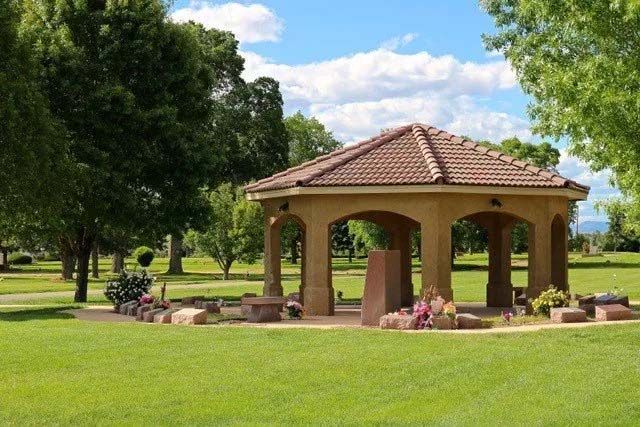 Gazebo in a sunny, green park with tiled roof, surrounded by landscaping, tombstones and flowers.