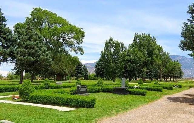 Cemetery with green grass, trees, and headstones under a blue sky. A dirt path runs through the scene.
