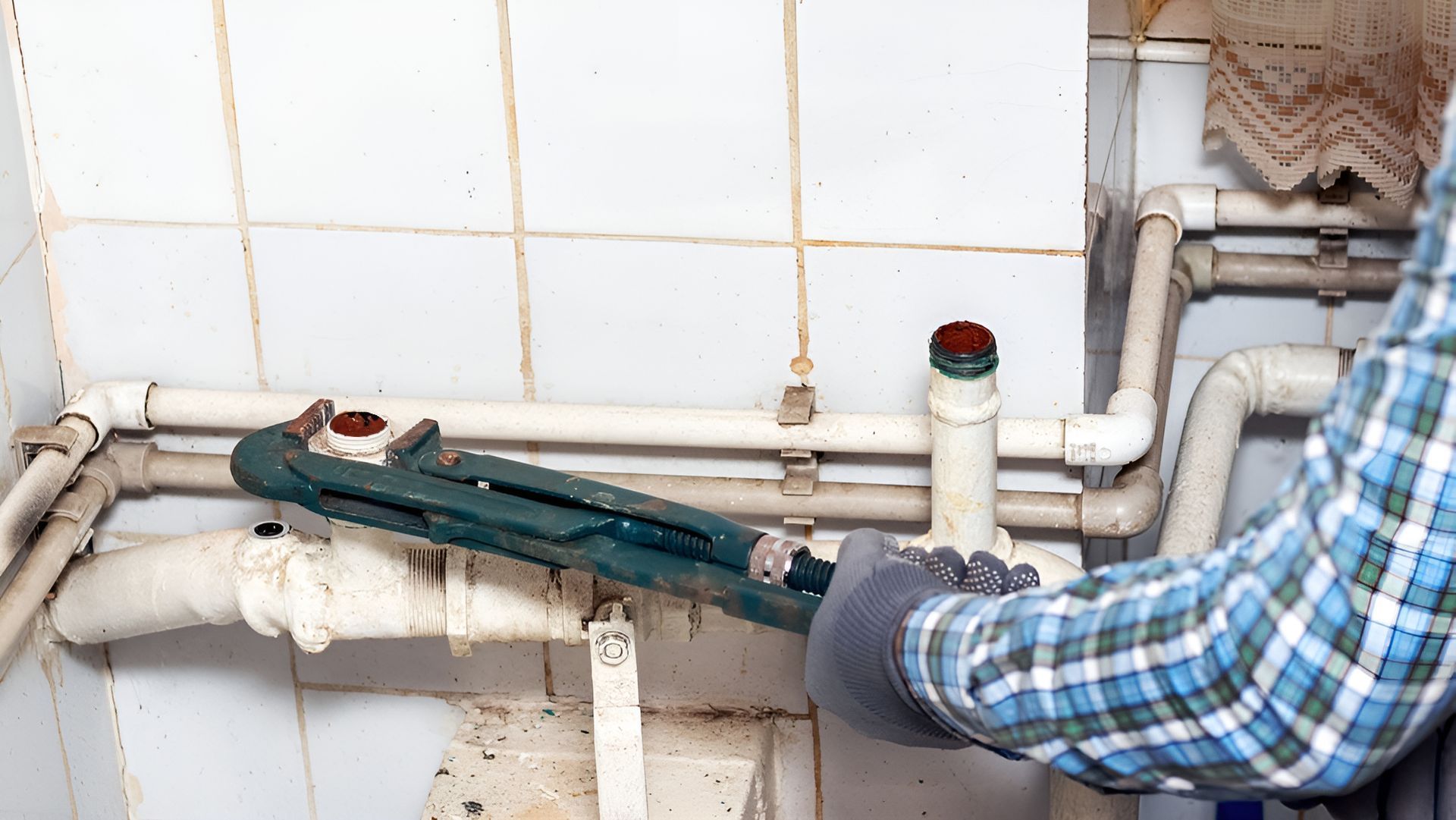 Plumber in a plaid shirt uses a wrench on white pipes in a utility room with white tile.