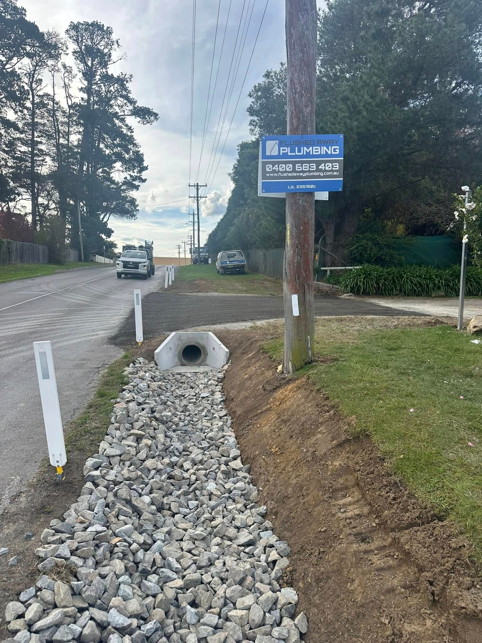 Gravel-lined drainage beside road with culvert, utility pole with sign. Flushed Away Plumbing in Bowral