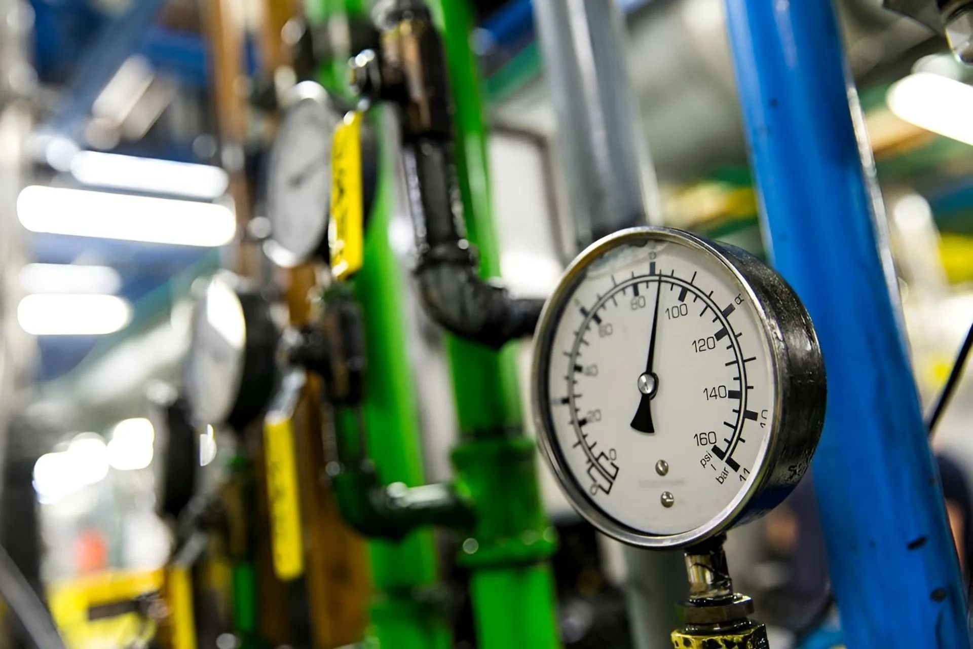 Pressure gauge on pipes, with green, black, and blue plumbing, indoors.