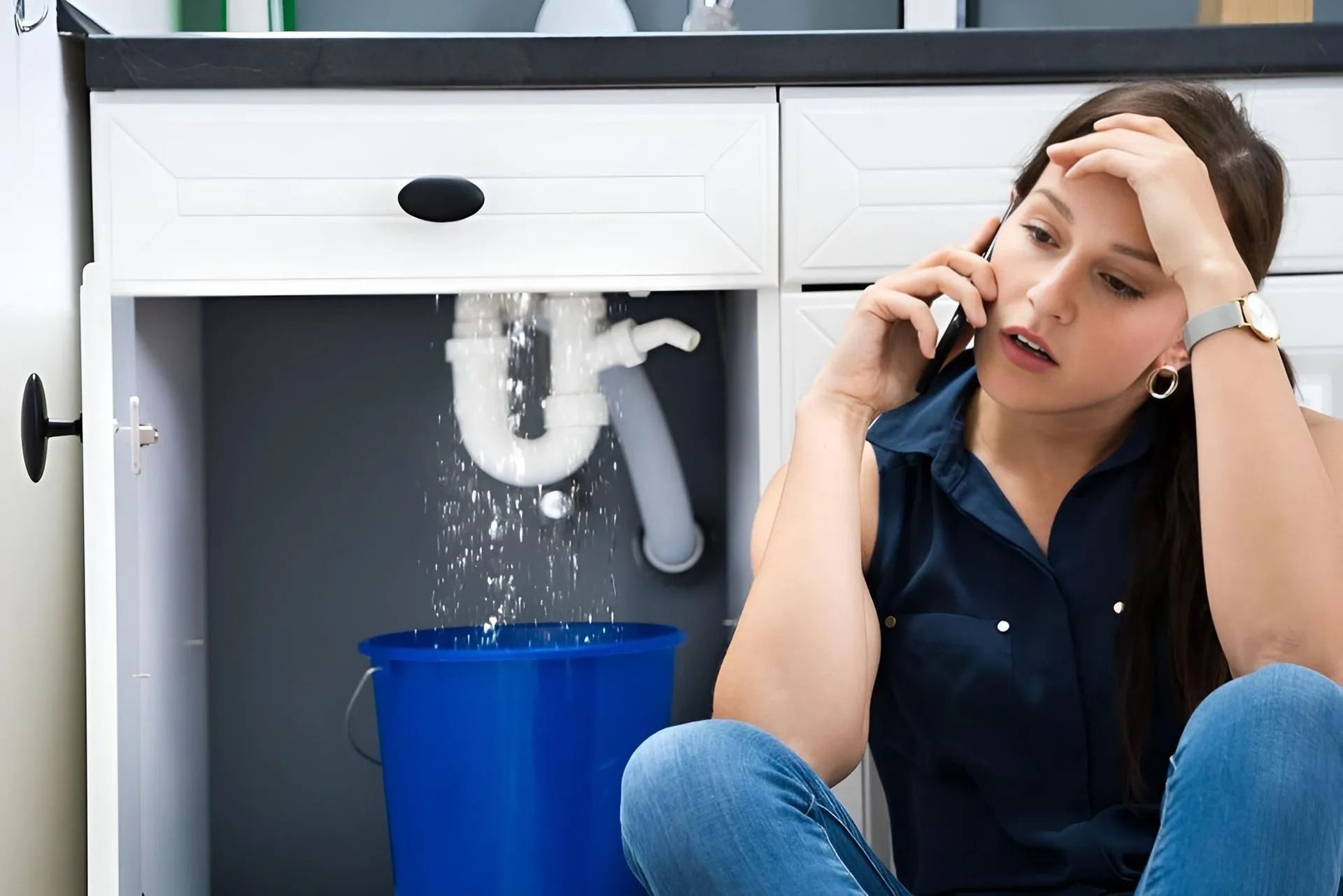 Woman on phone, distressed, sits near a leaking sink. Water spills into a blue bucket.