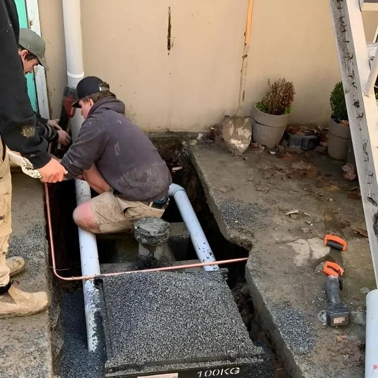 Two workers installing plumbing pipes in a trench outside, with a black utility box.