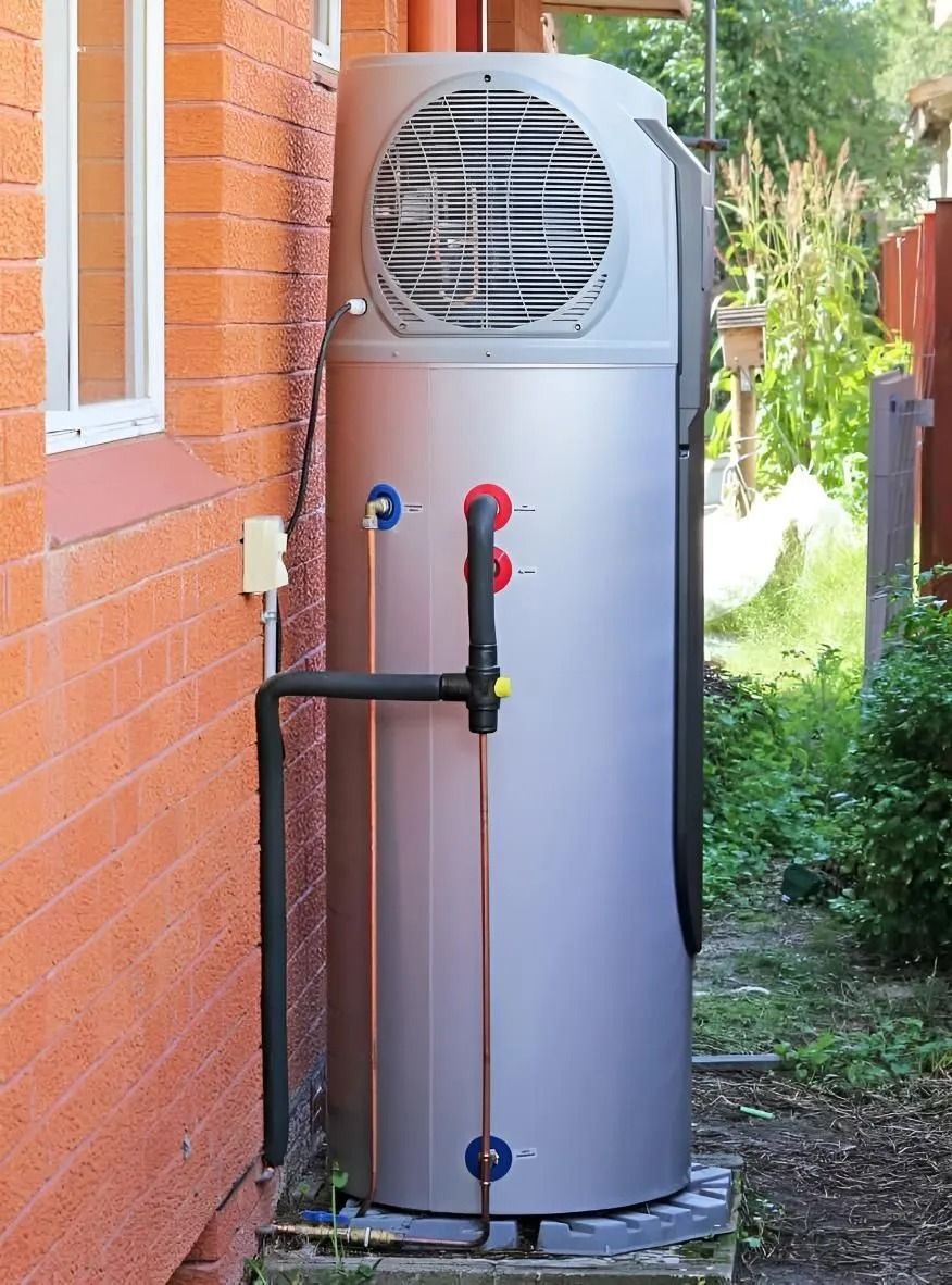 A tall, silver heat pump water heater installed outside against a red brick wall and foliage.