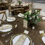 Outdoor restaurant table set with gold-rimmed plates, white napkins, glasses, and a floral centerpiece on a wooden deck