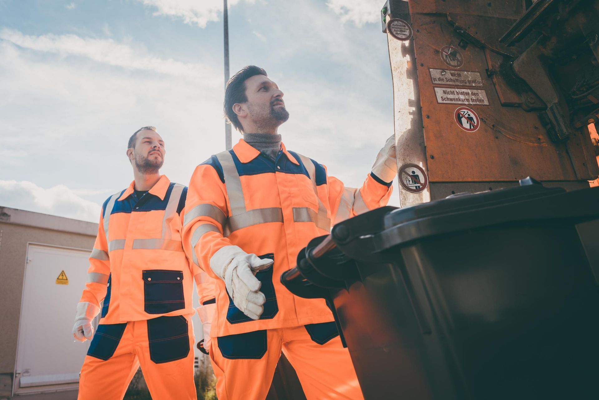 Two waste workers in orange and navy uniforms emptying a trash bin into a truck, under a cloudy sky.