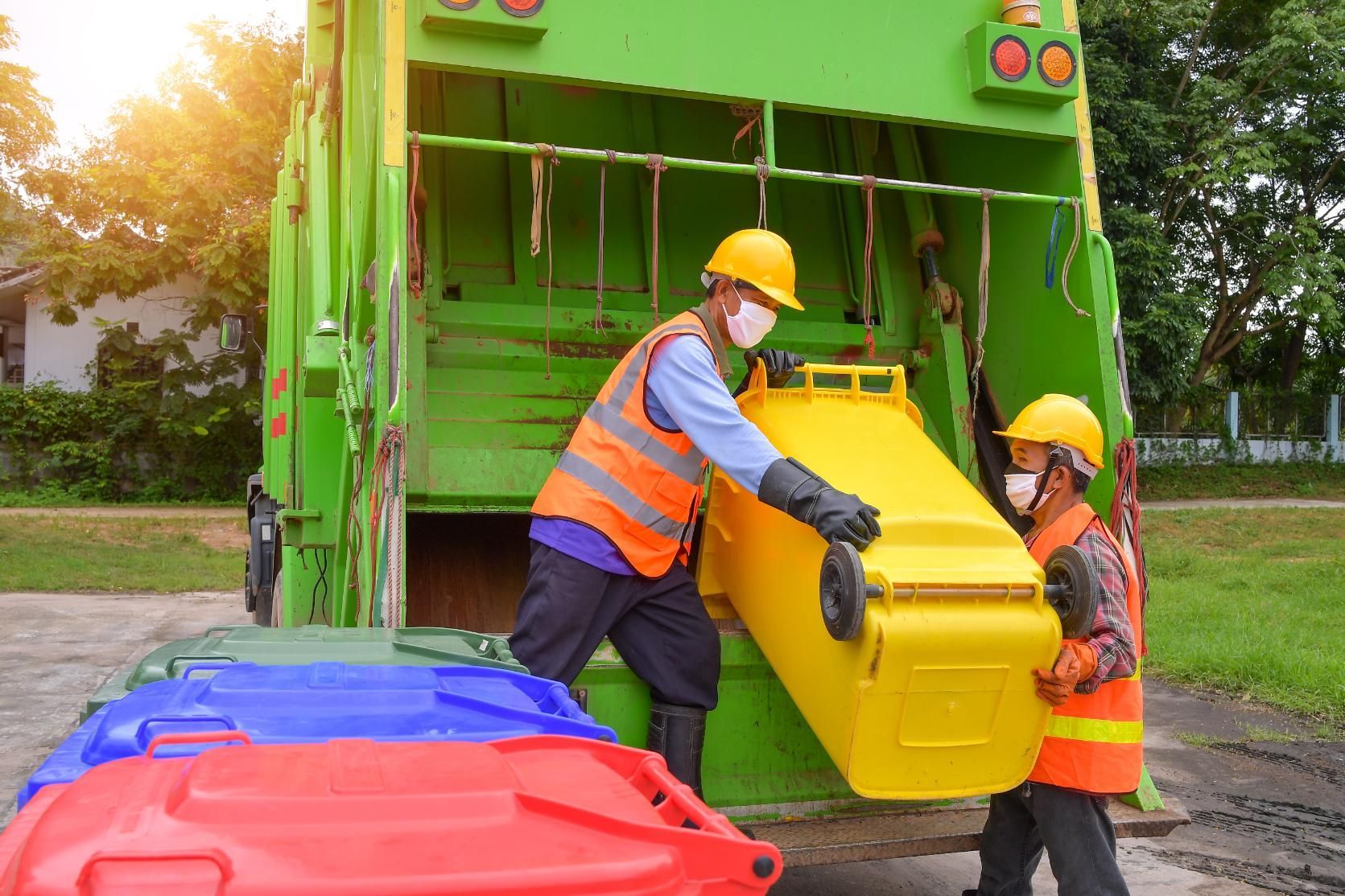 Two sanitation workers emptying a yellow recycling bin into a green garbage truck.