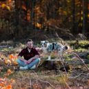Mann sitzt im Schneidersitz am Boden, daneben sein Hund auf einem Baumstamm