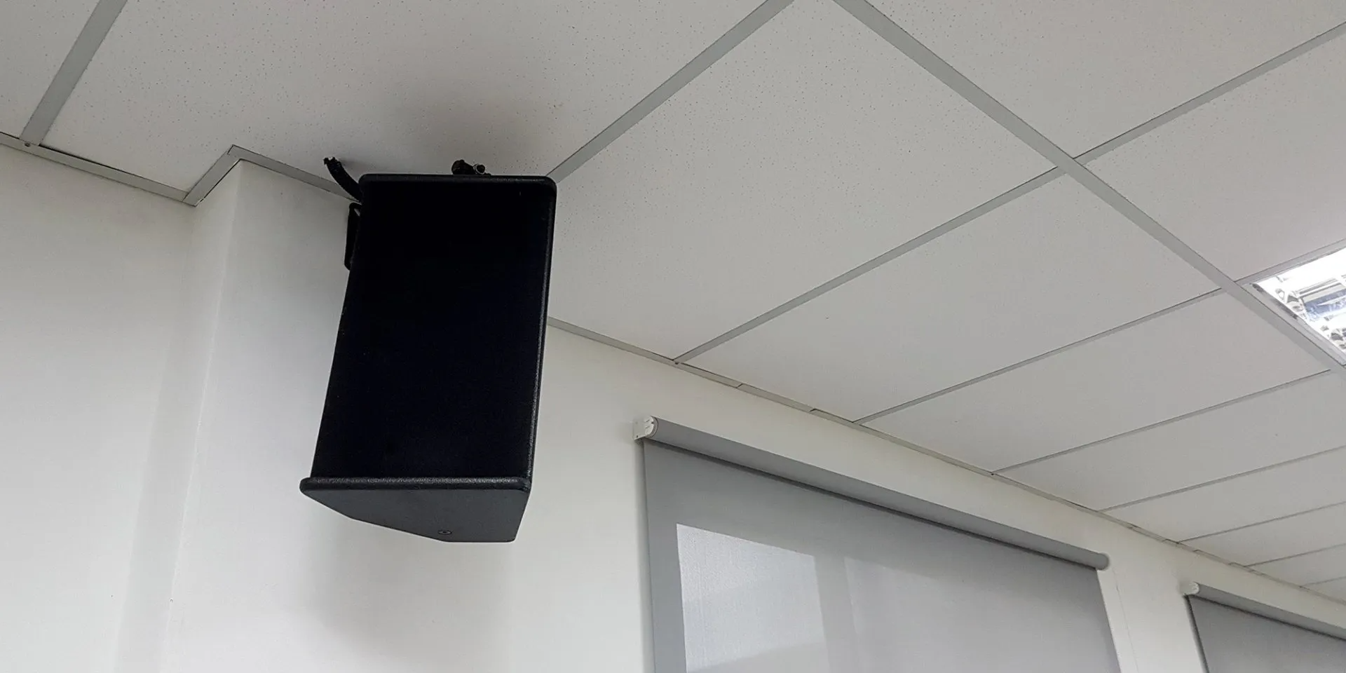 Black wall-mounted speaker near a ceiling tile and doorway in a white room