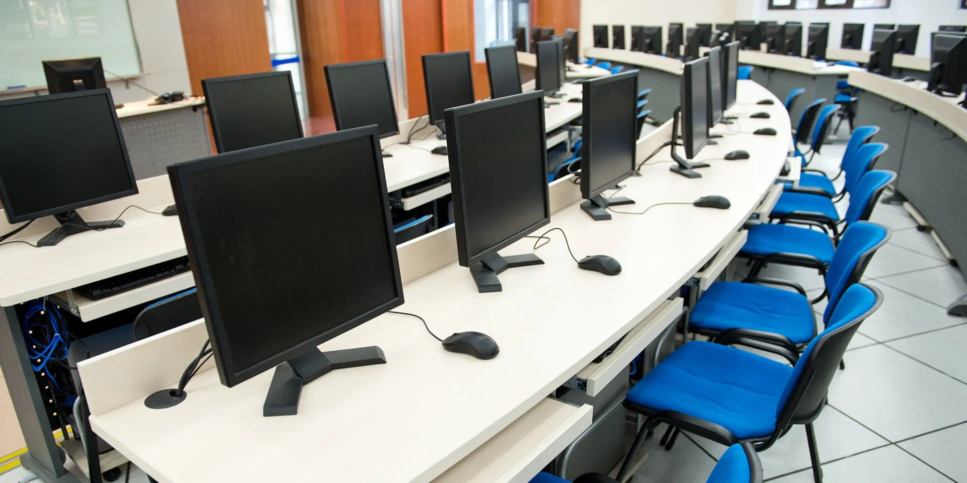Empty computer lab with rows of desktop monitors and blue chairs around curved desks.