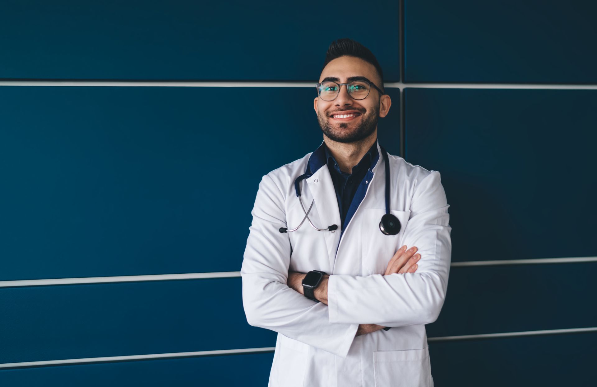 Doctor with stethoscope, arms crossed, smiling in front of a blue wall.