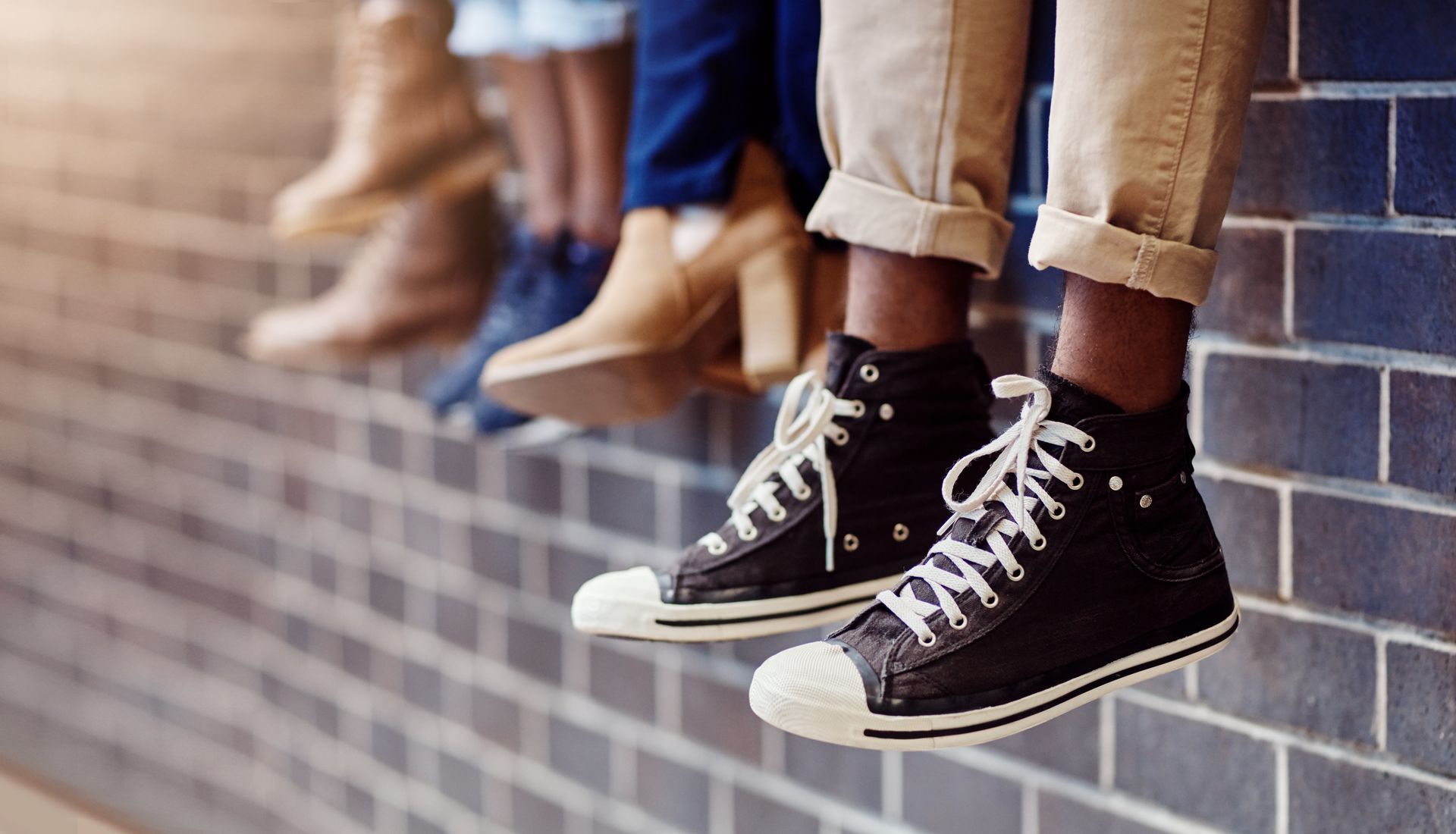 Several pairs of feet wearing different shoes hang over a brick wall.