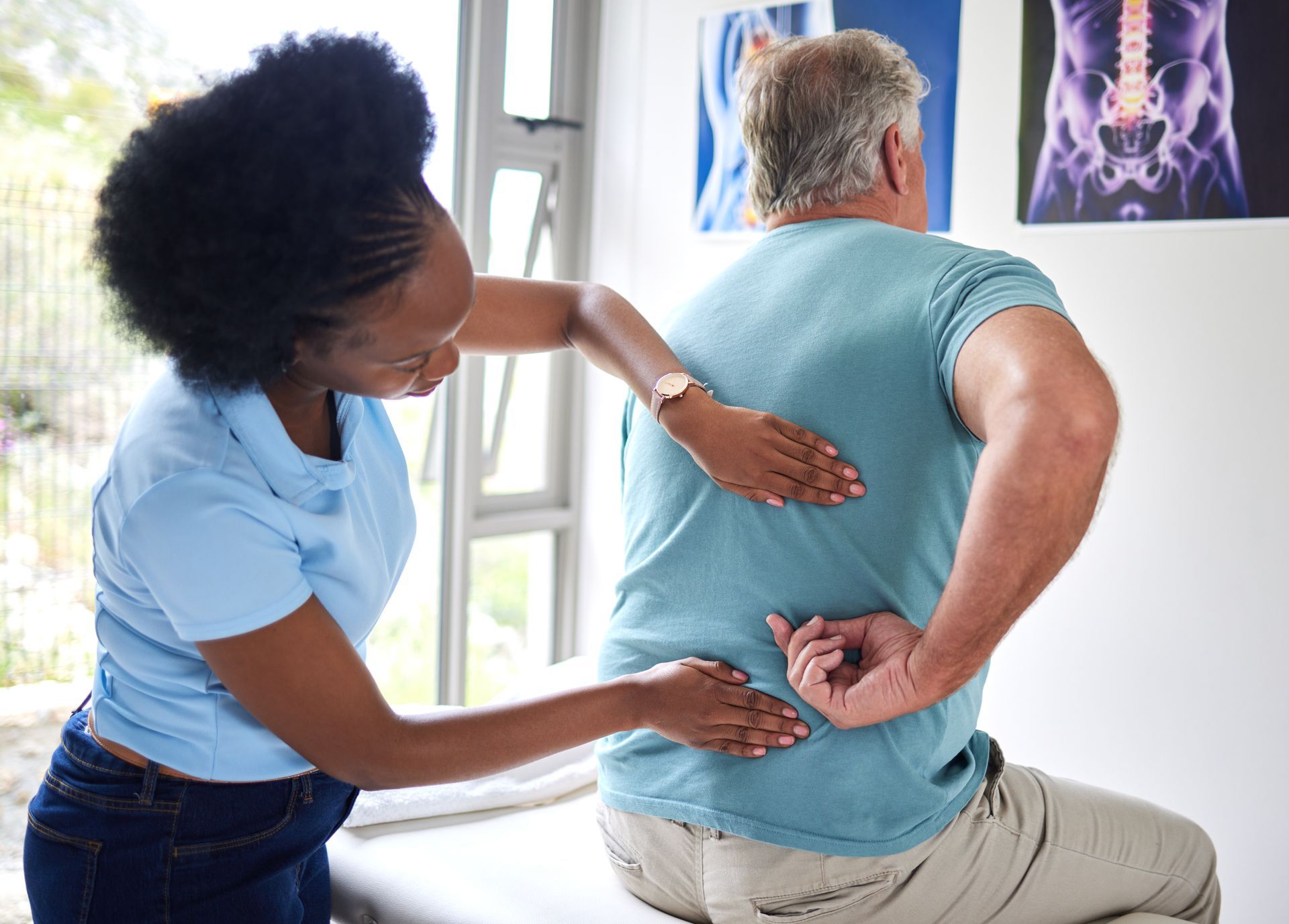 Woman examining man's back in a light-filled medical office, hands on his lower back. X-ray images on wall.