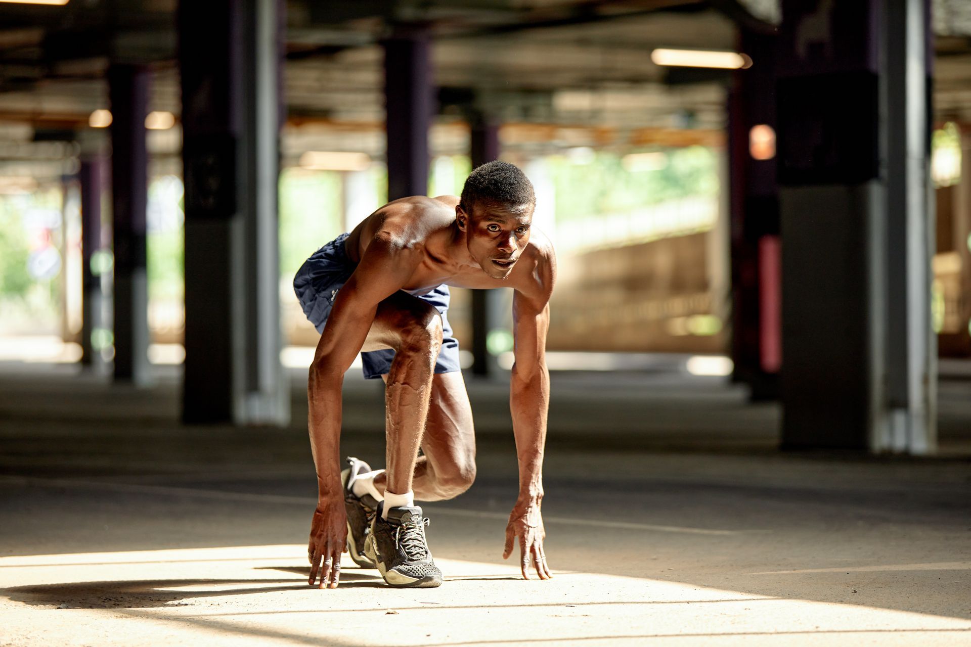 Runner in a low starting position under a concrete structure; sunlight.