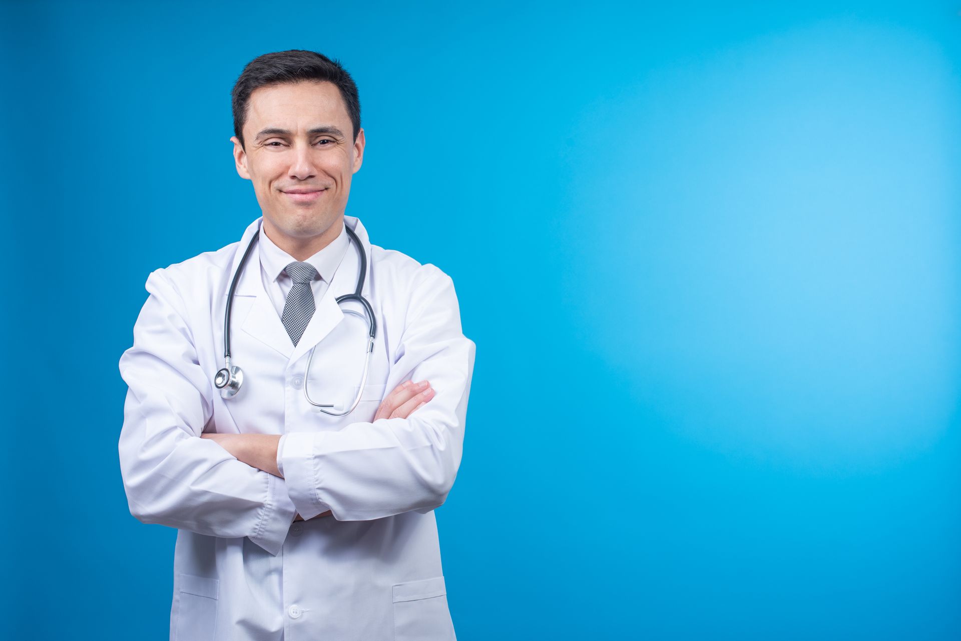 Doctor with a stethoscope, arms crossed, smiles against a blue background.