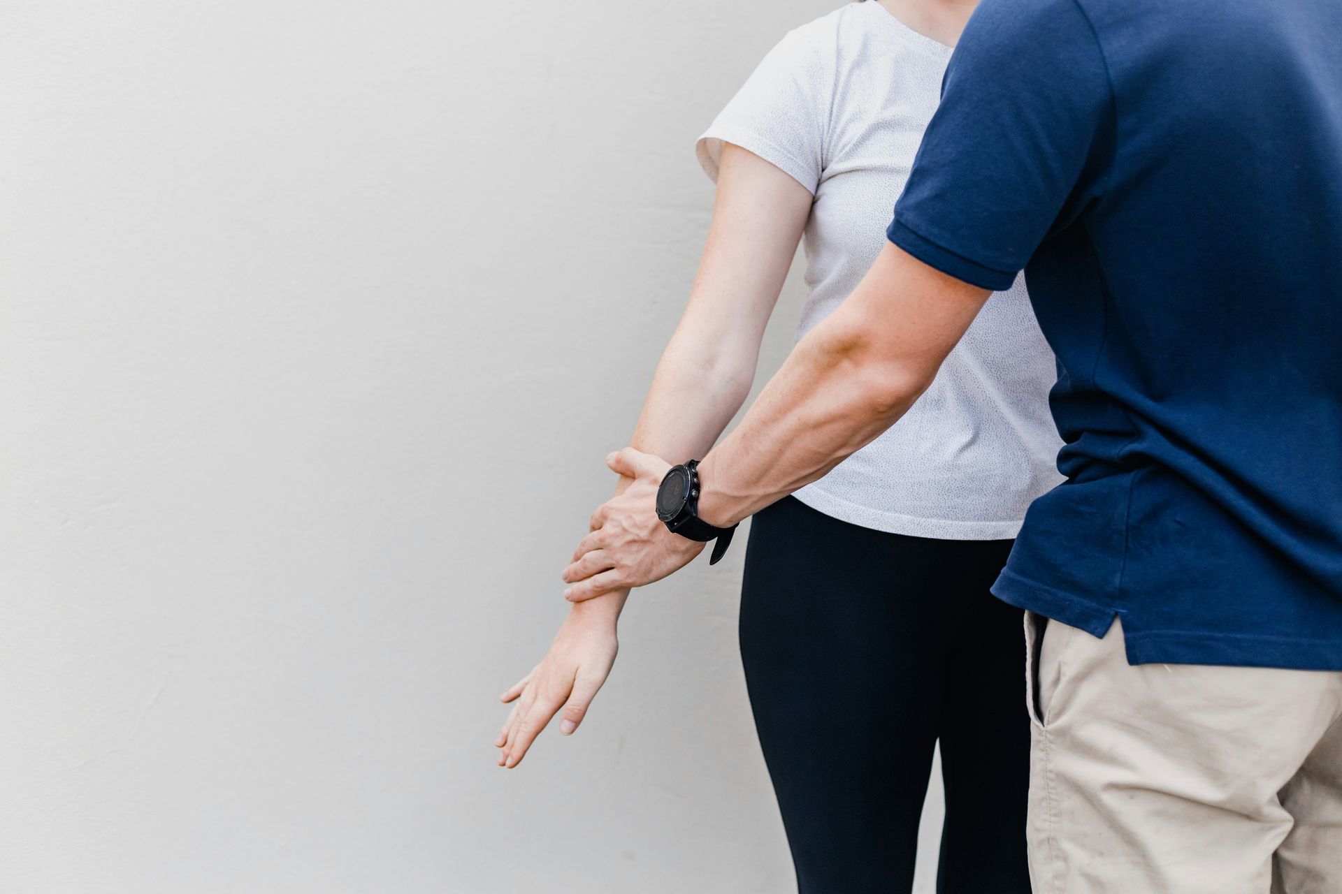 Person supporting another person's arm, likely for physical therapy or assessment, against a plain wall.