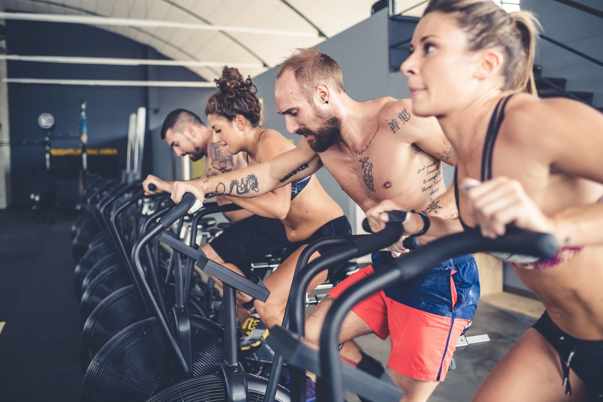 People exercising on fan bikes in a gym, faces showing exertion.
