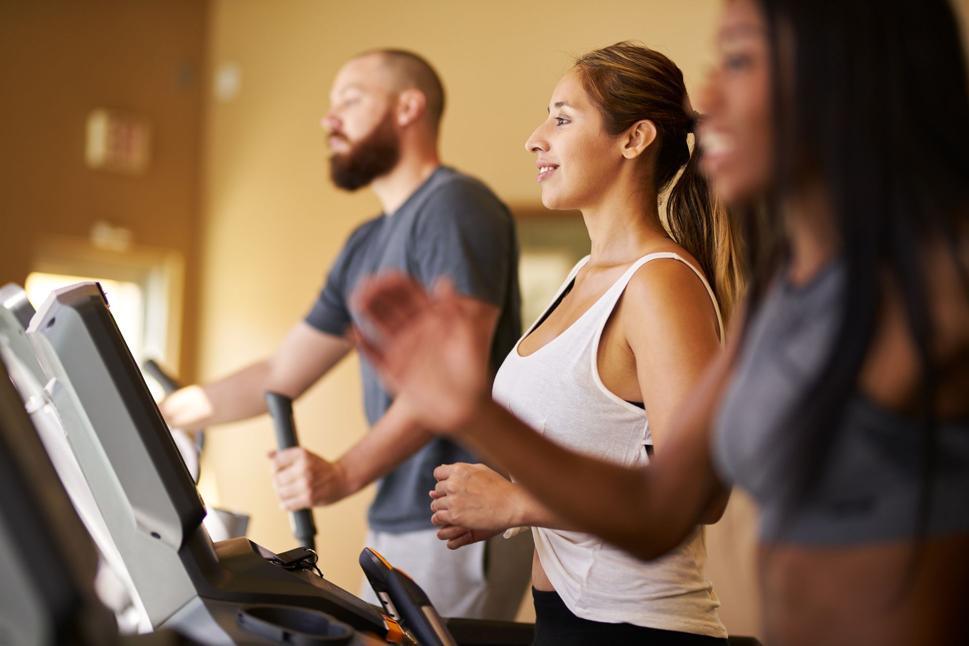 People exercising on treadmills in a gym. A woman in a white tank smiles, another in gray. A man is behind her.