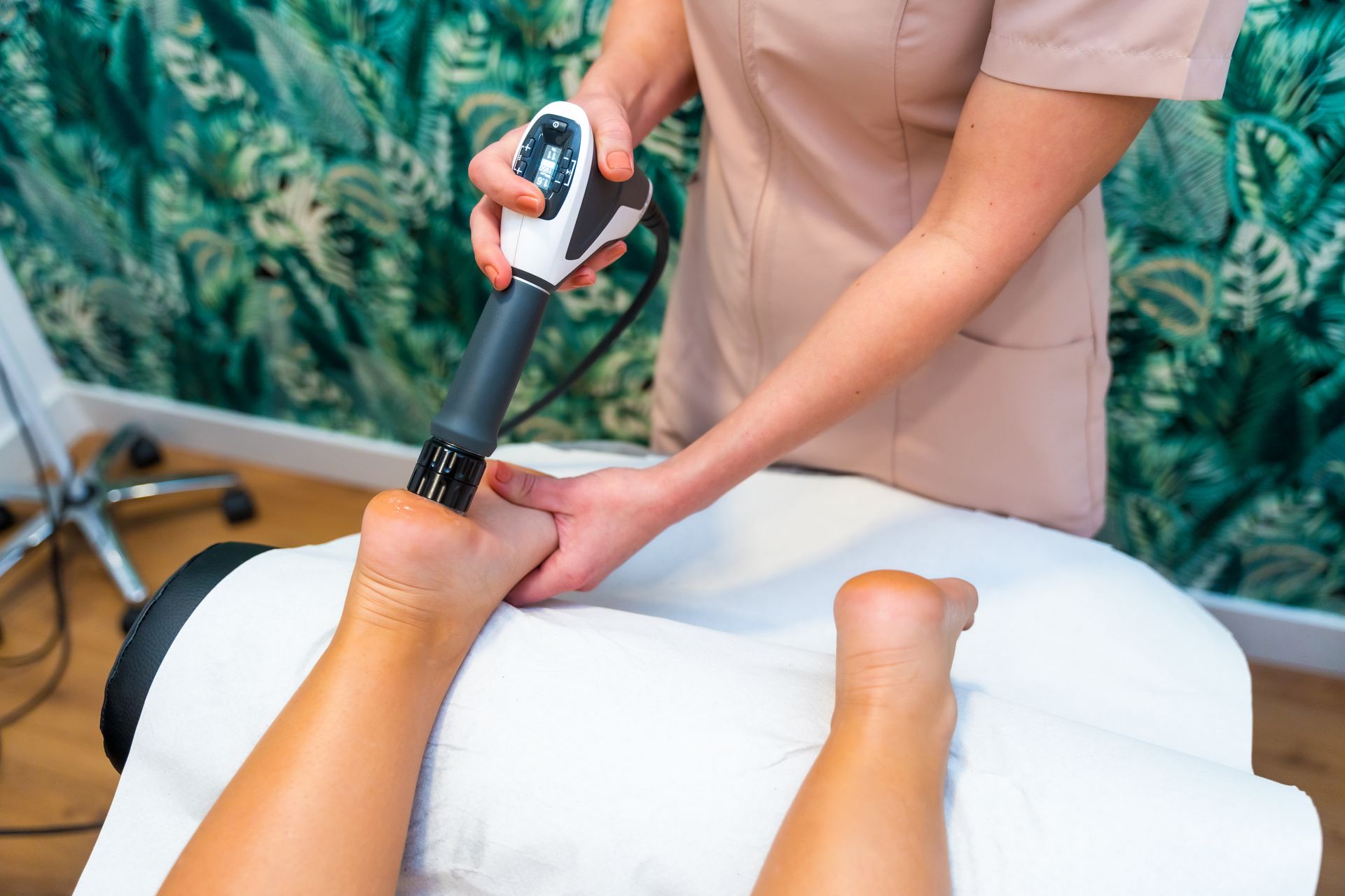 Chiropractor using a shockwave therapydevice on a patient's foot, indoors, white bed, green wall.