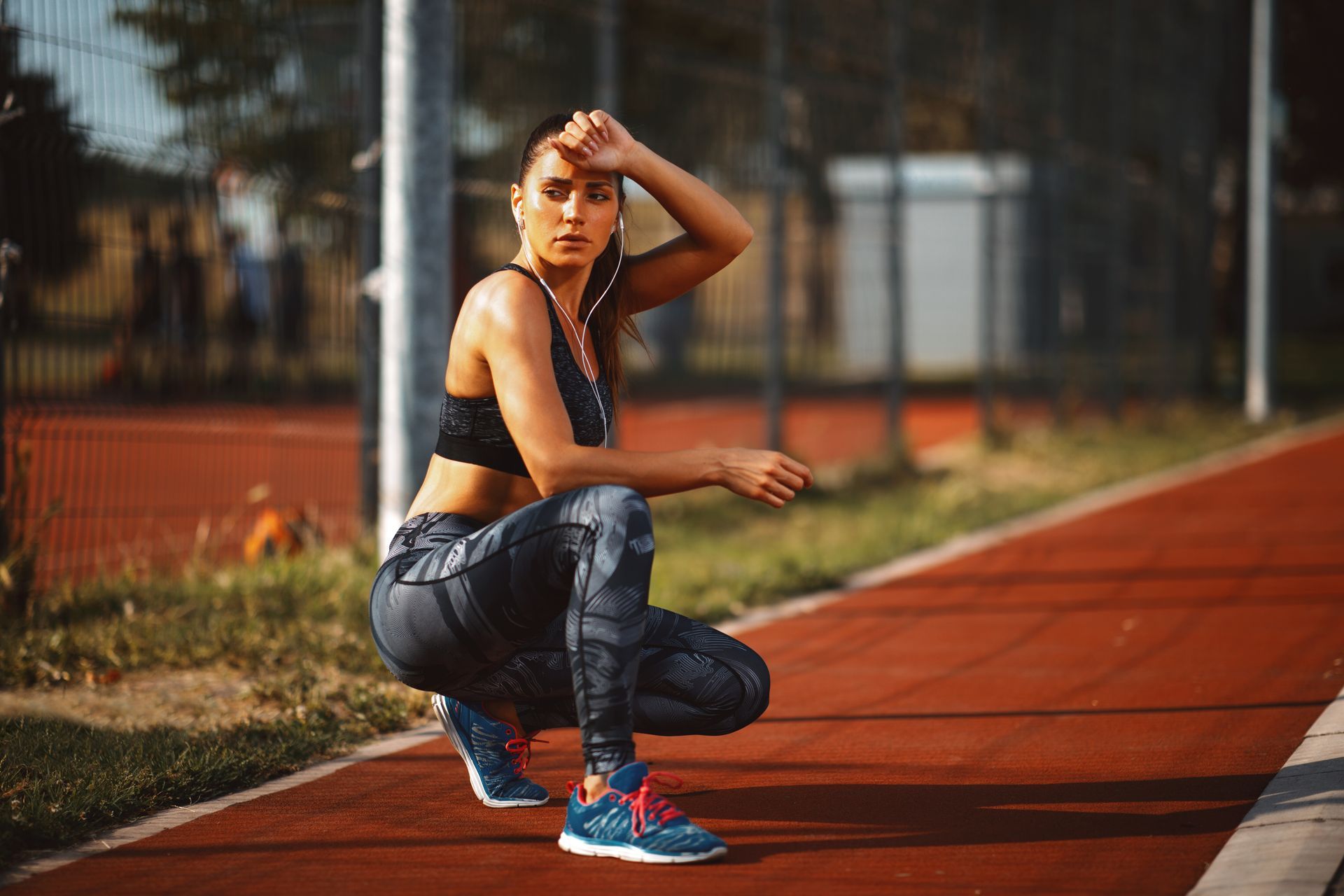 Woman in workout clothes rests on track, wiping forehead, in sunlight.