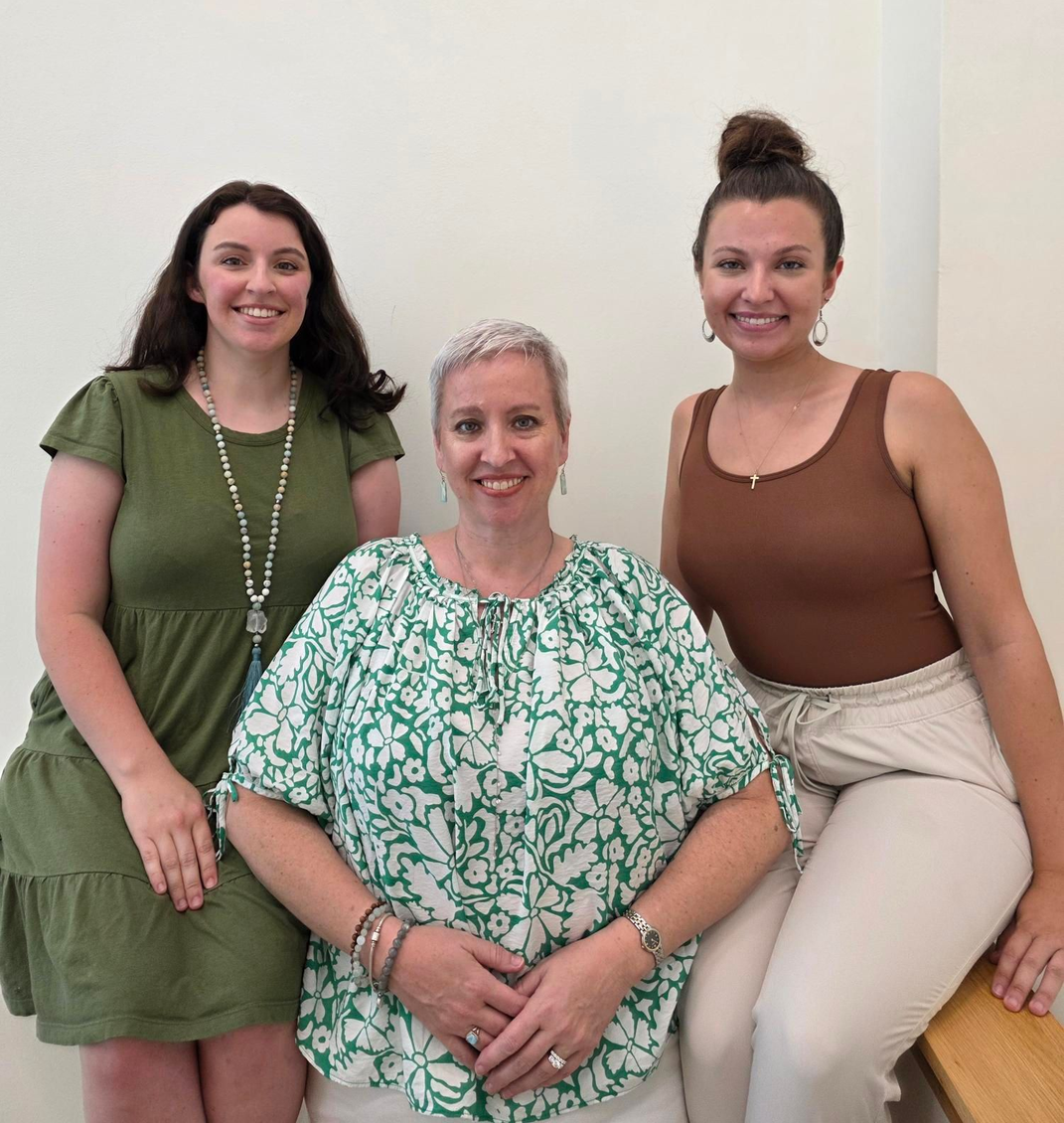 Three women smiling and sitting together indoors against a plain wall.