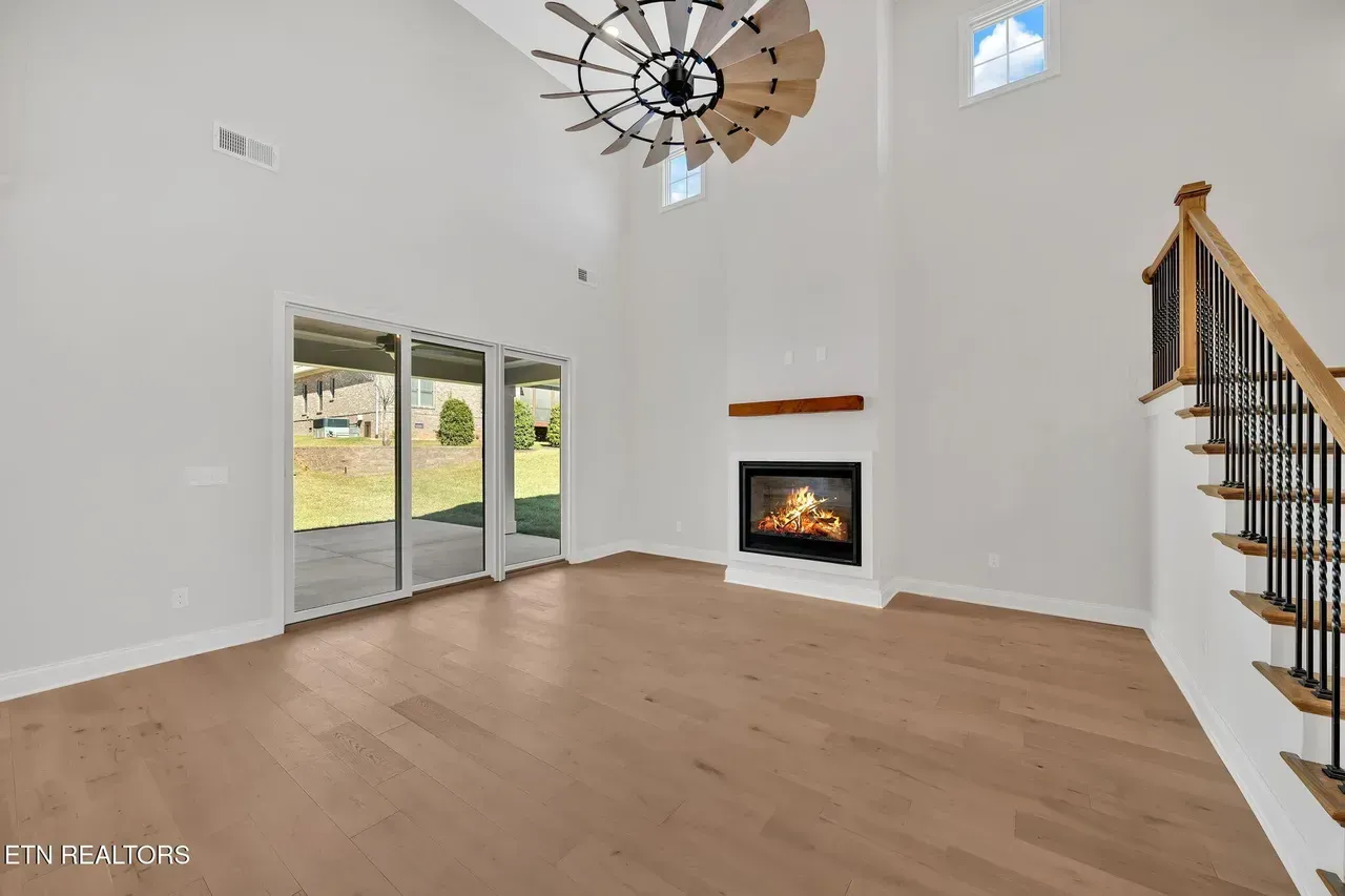 Empty living room with fireplace, sliding doors, and staircase. Light wood floors, white walls, and a large ceiling fan.