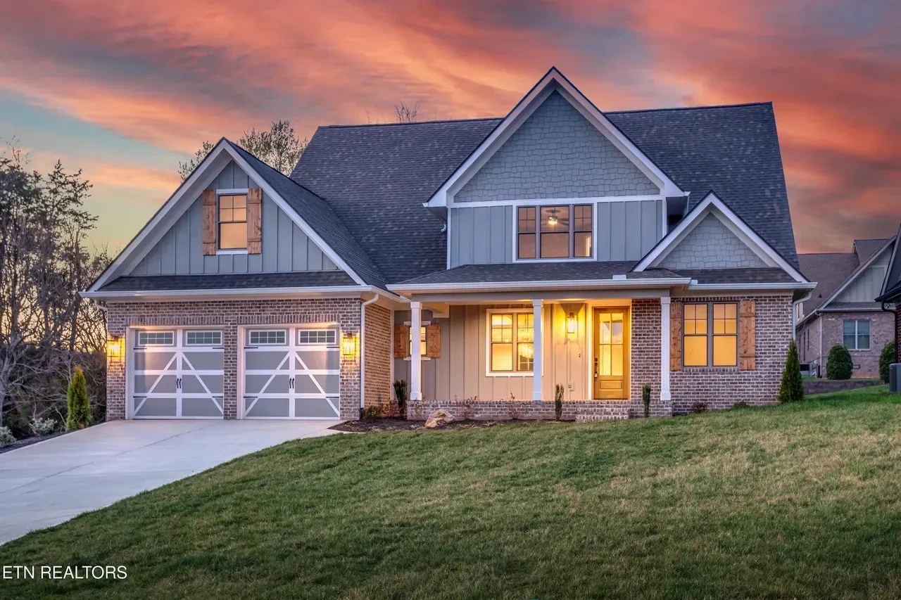 House with light blue siding, a covered porch, and a two-car garage at dusk, set on a green lawn with a sunset sky.