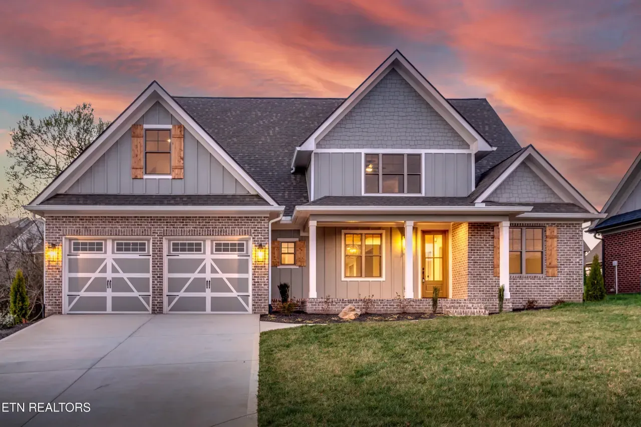 House with gray and brick facade, two-car garage, and porch; set against a sunset sky.
