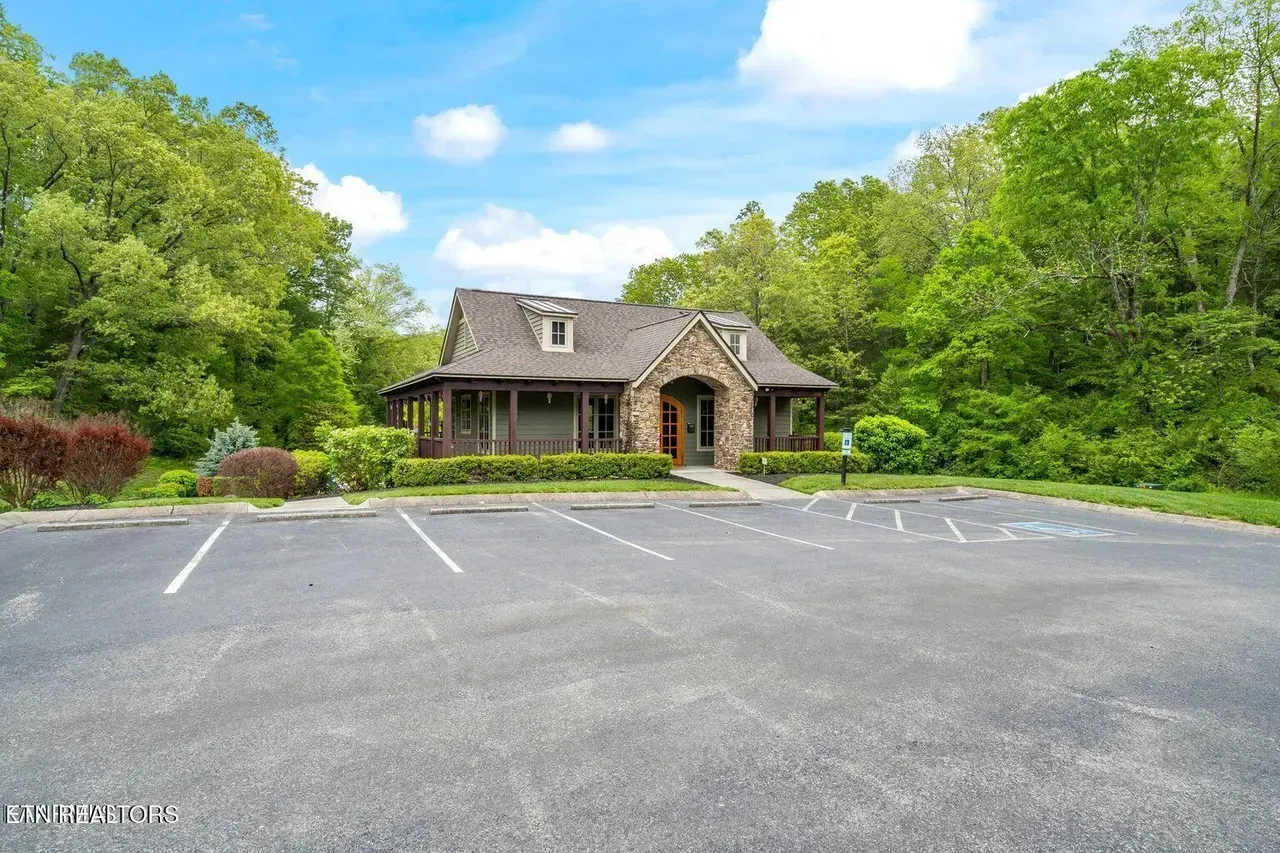 Stone cottage with a porch, surrounded by trees, and a parking lot in front. Blue sky.