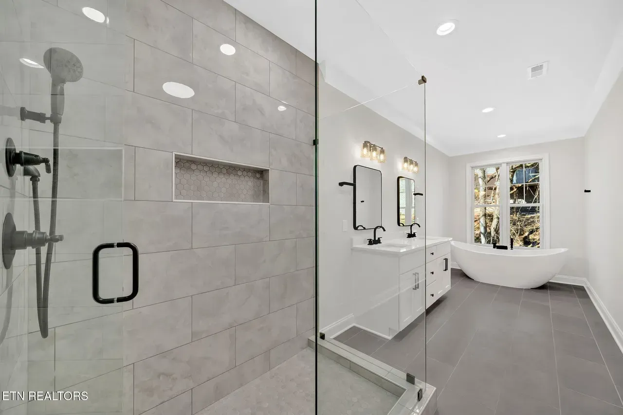 Modern bathroom with glass shower, double sinks, and a freestanding tub. Gray tiles and white walls.