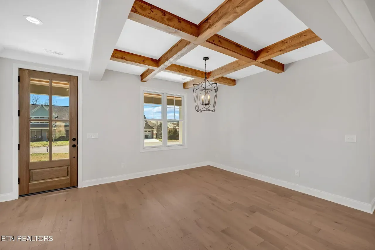 Empty dining room with wood floors, light gray walls, wooden ceiling beams, chandelier, and a front door.