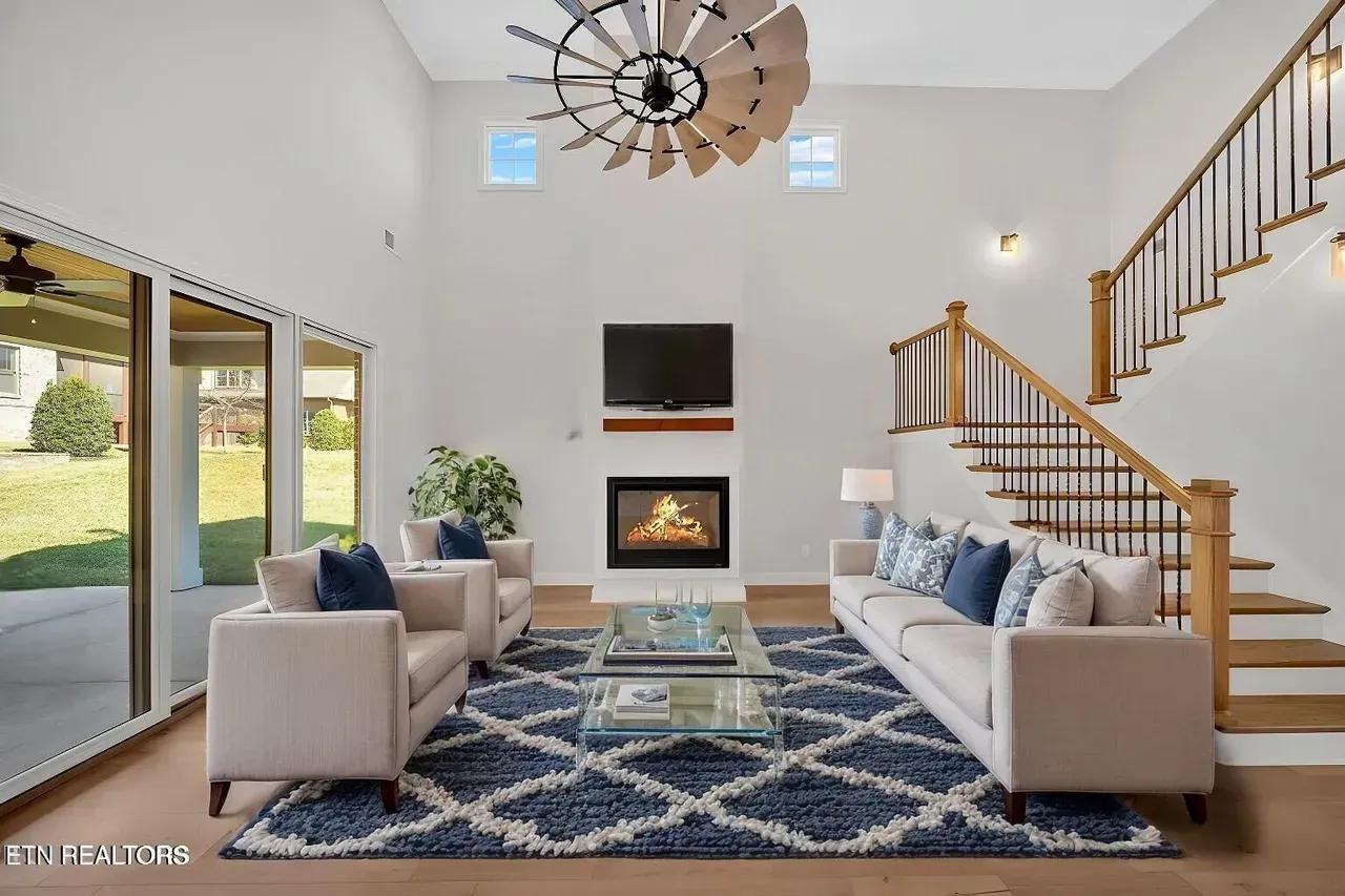 Modern living room with fireplace, stairs, blue rug, and large windows overlooking a yard.