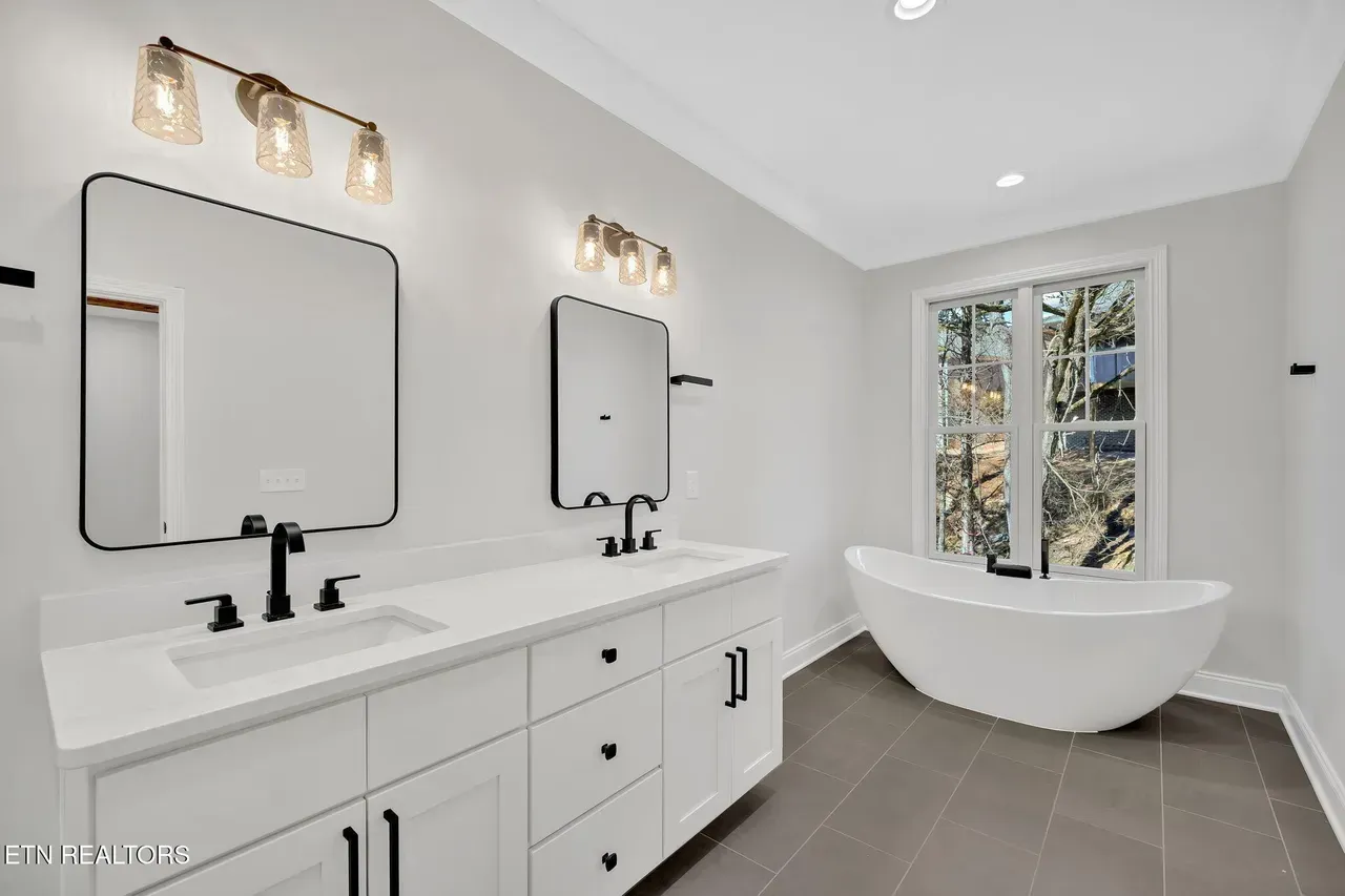 Modern white bathroom with double vanity, bathtub by a window, and gray tiled floor.