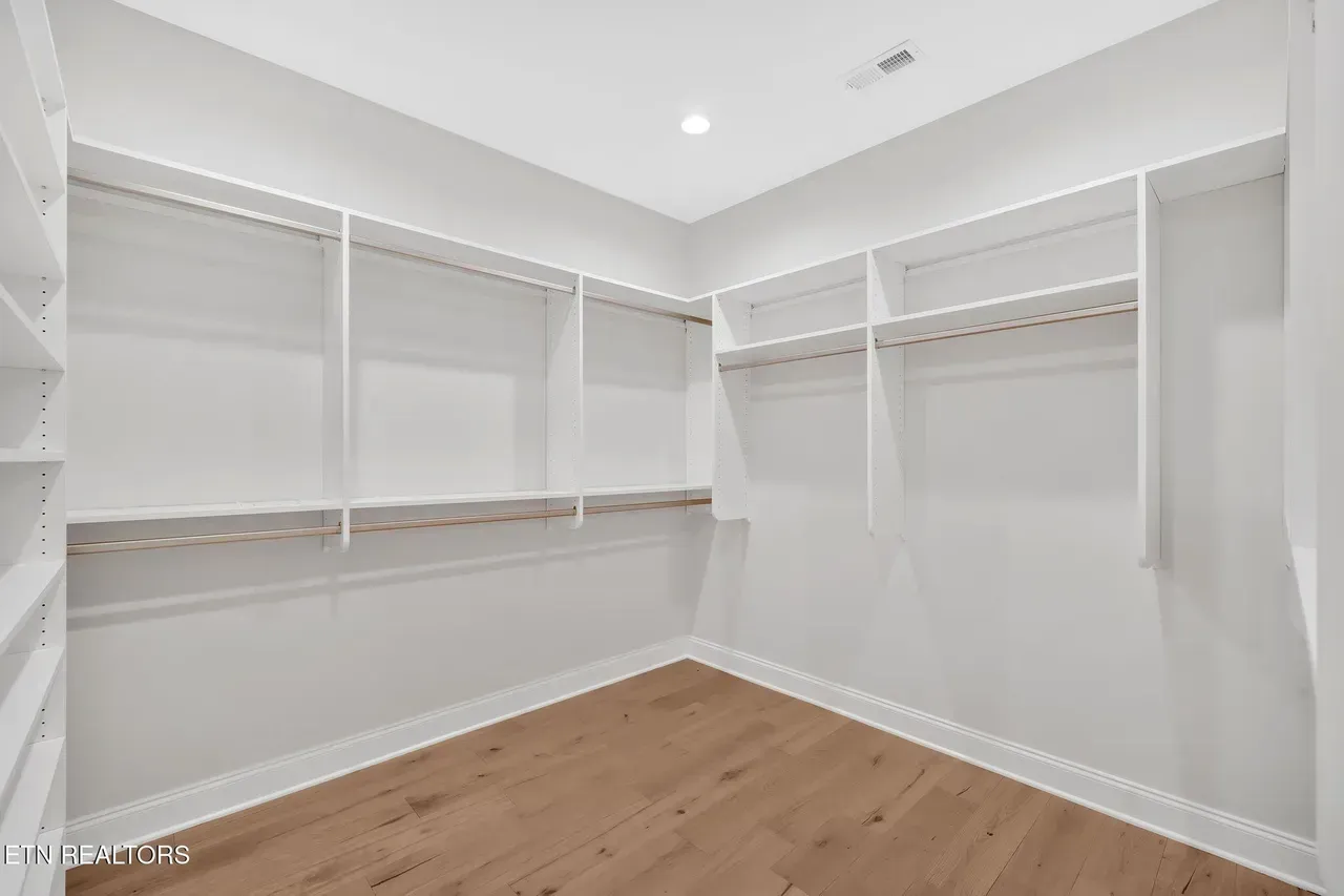 Empty white walk-in closet with shelves and hanging rods, wooden floor, and overhead light.