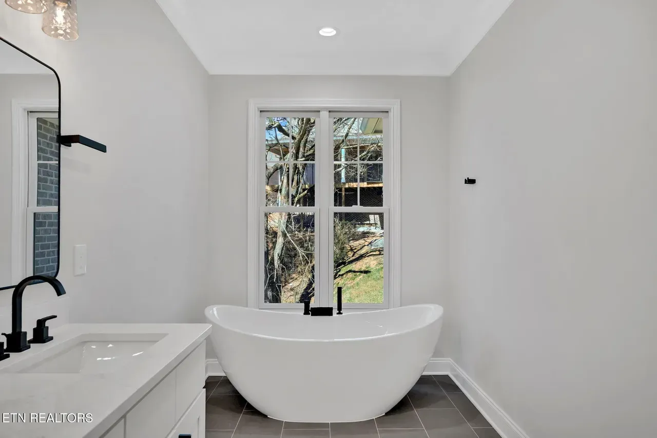 Modern bathroom with white freestanding tub, black fixtures, and a window overlooking greenery.