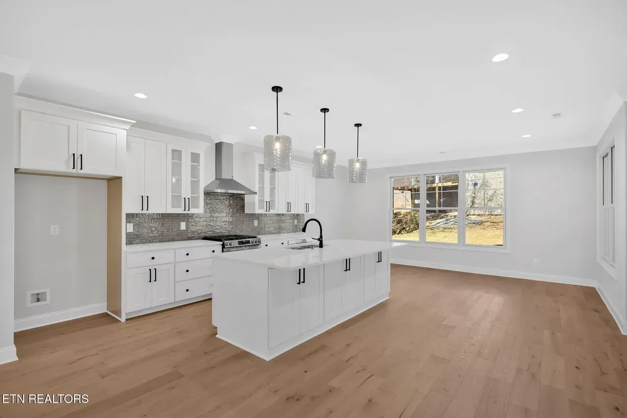 White kitchen with island and pendant lights; stainless steel appliances, wood floor.