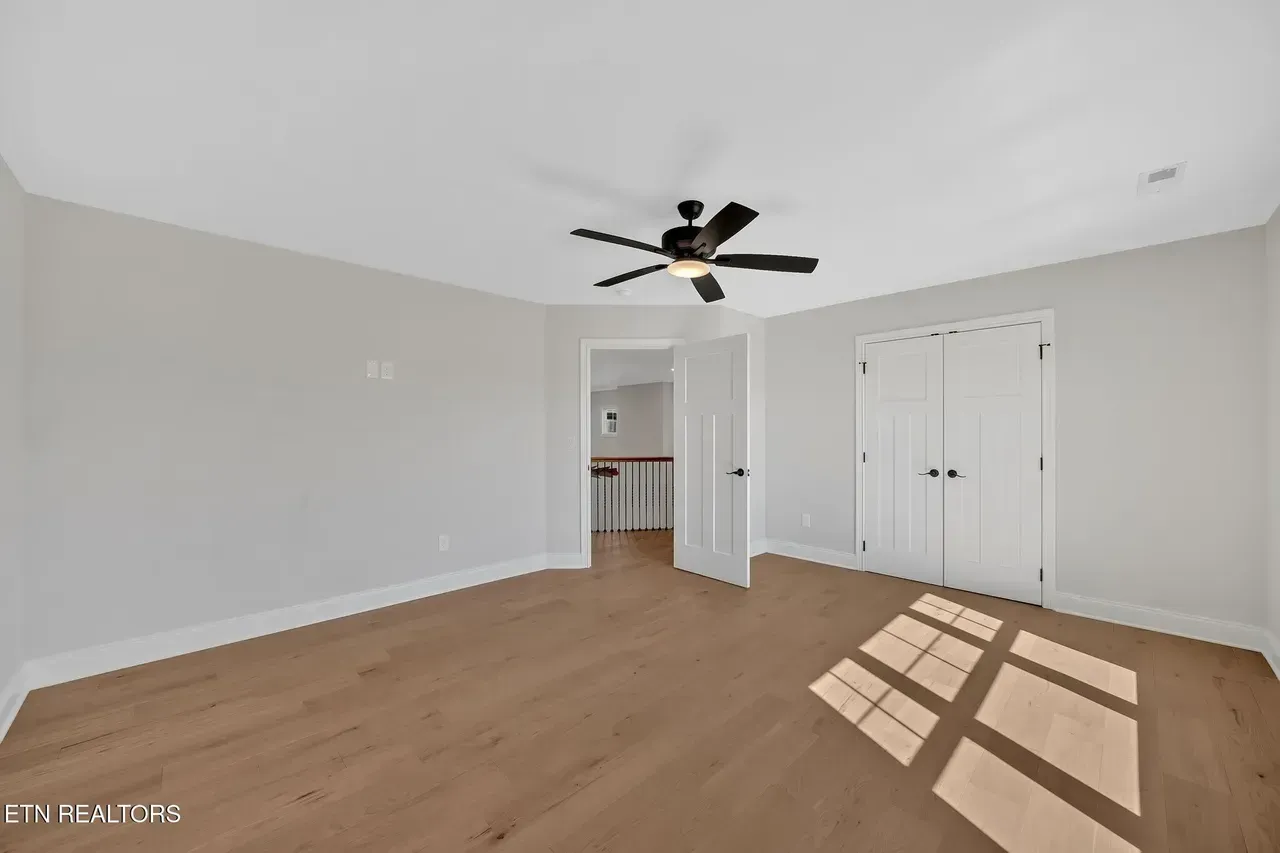Empty room with hardwood floors, white walls, closet, open doorway to hallway, ceiling fan.