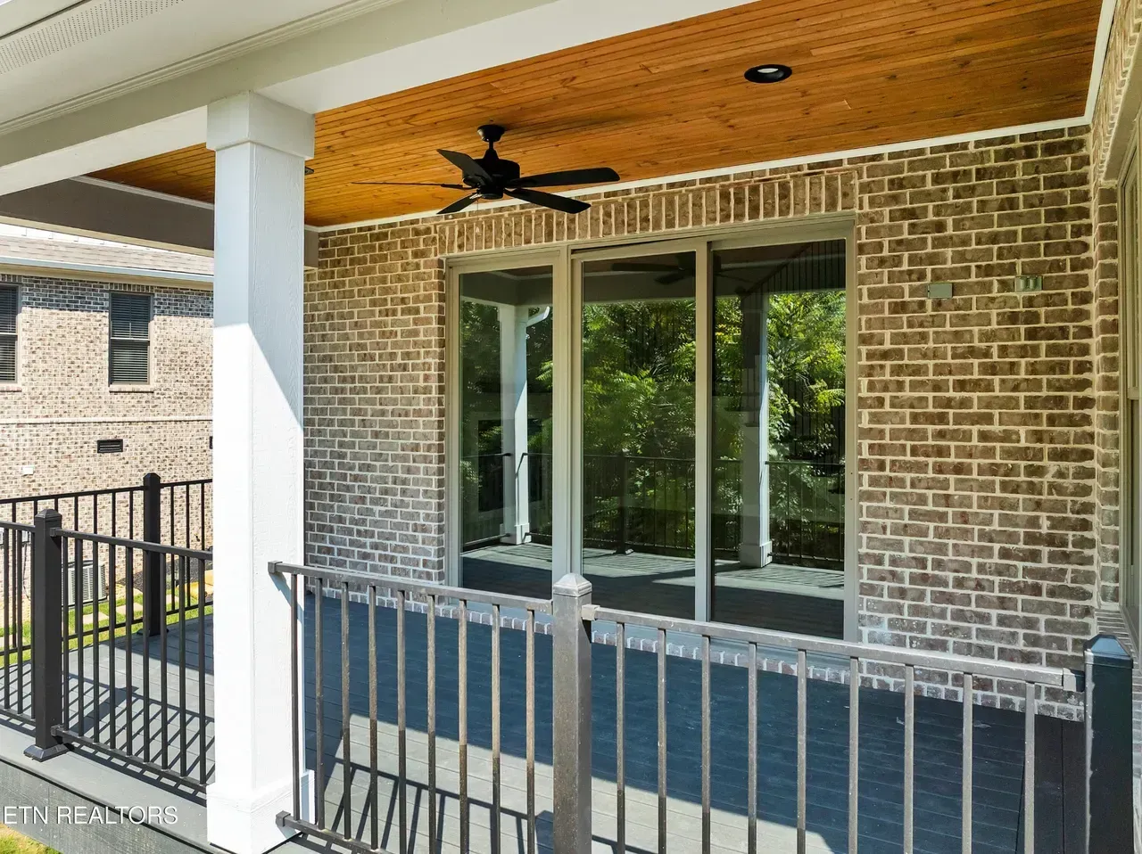 Brick porch with black railing, large windows, and a ceiling fan, with a view of greenery.