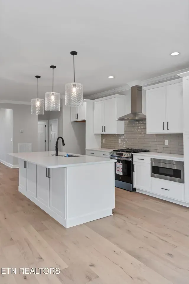 White kitchen with island, pendant lights, stainless appliances, and light wood floors.