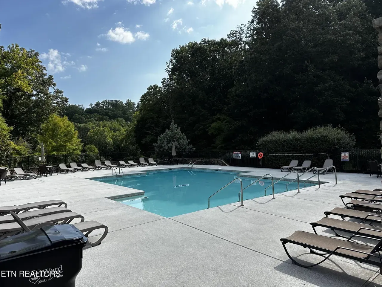 Outdoor swimming pool surrounded by lounge chairs, trees, and a blue sky.