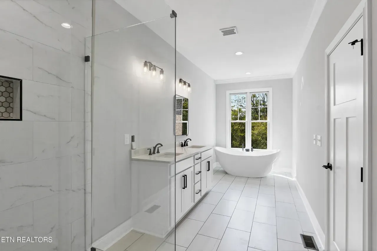 Modern white bathroom with double vanity, shower, and freestanding tub near a window.