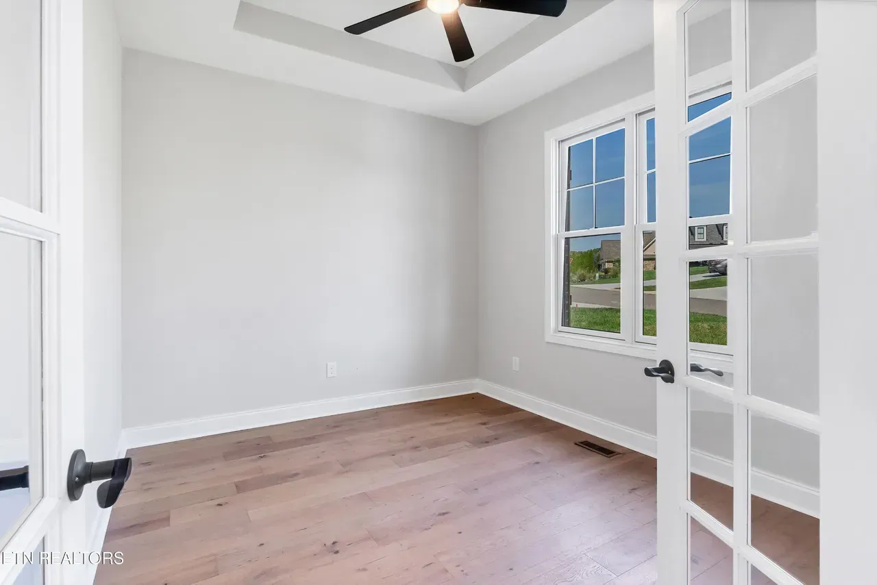 Empty room with light gray walls, wooden floor, and large window. Open French doors on the right.