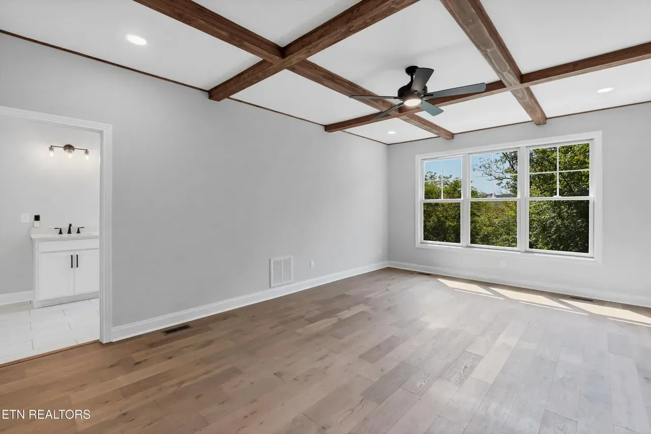 Empty room with wood floors, large window, coffered ceiling, and doorway to bathroom.