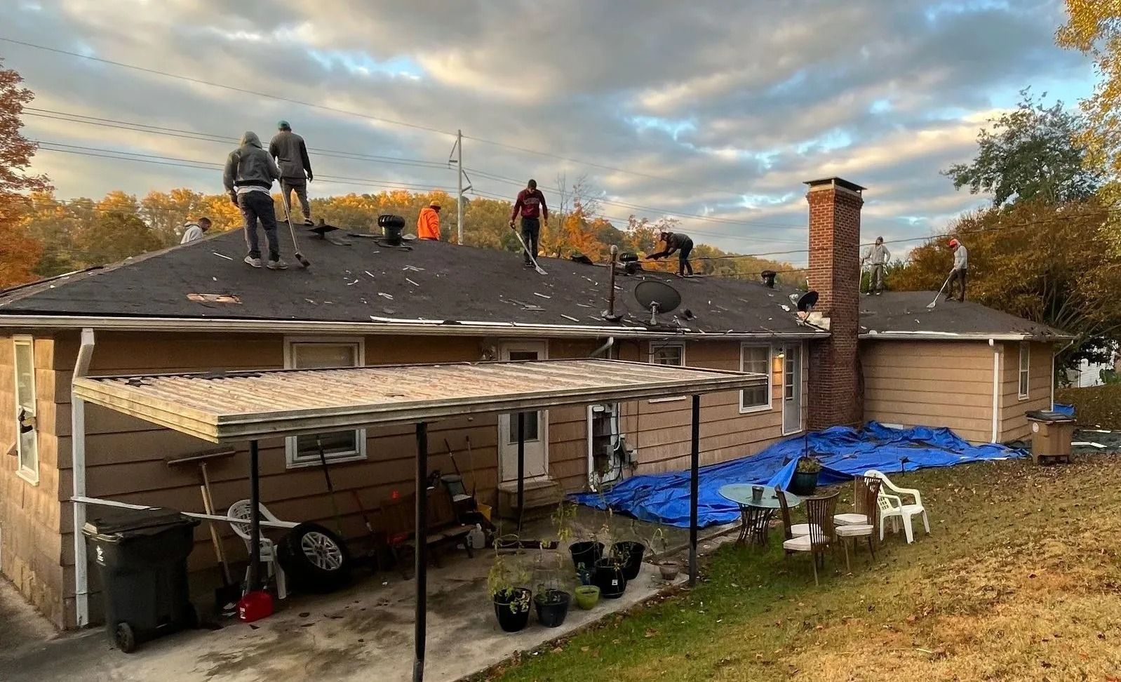 Workers on a roof, removing shingles. A chimney and carport are visible. Overcast sky, brown house.