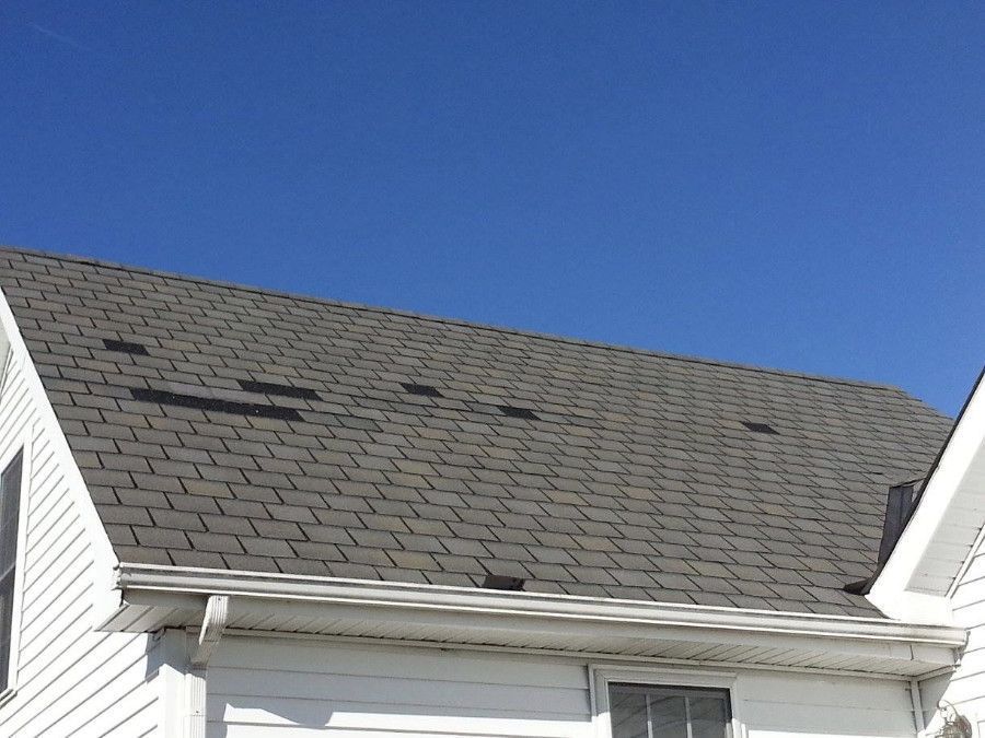 Gray shingled roof of a white house with missing shingles against a clear blue sky.