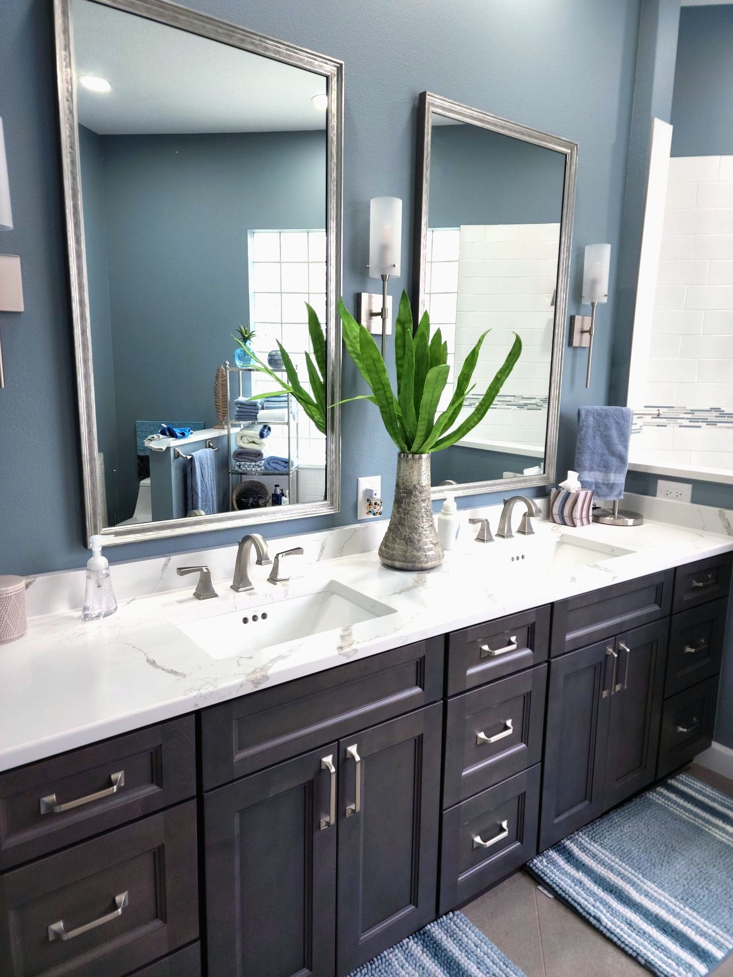 Bathroom with dark gray vanity, white countertop, two mirrors, blue walls, and a vase with green plant.